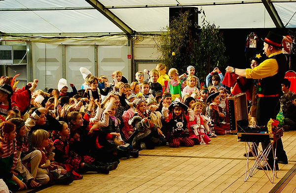 Kinderzauberer Markus Poétes steht auf der Bühne bei einer Kindersitzung und begeistert eine große Gruppe Kinder, viele Hände sind begeistert in der Luft