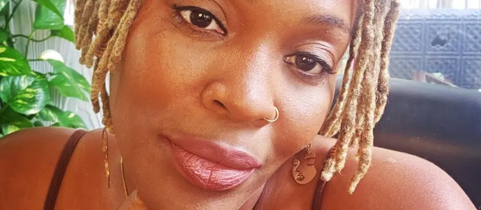 Woman with locs, wearing a brown tank top, gazes calmly. Hand on chin, sitting indoors. Plants and macramé in background. Warm tones.