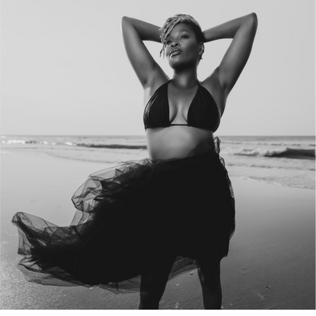 Woman posing confidently on a beach in a black bikini top and flowing skirt. Overcast sky and gentle waves in the background. Black and white image.
