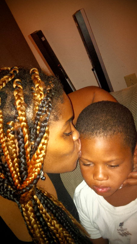 Woman with braided hair kisses boy on cheek. Boy looks upset. Indoor setting with neutral background. Emotional warmth contrasts mood.