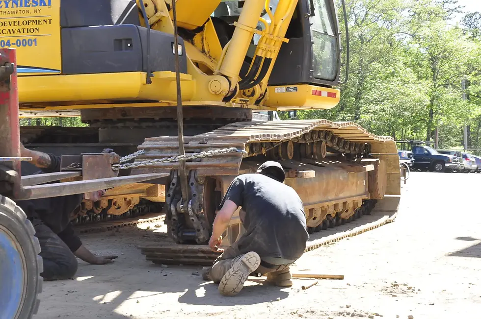 Worker kneeling beside yellow excavator, repairing its track. Sunlit outdoor setting with trees and vehicles in the background in Fort Lauderdale, FL.