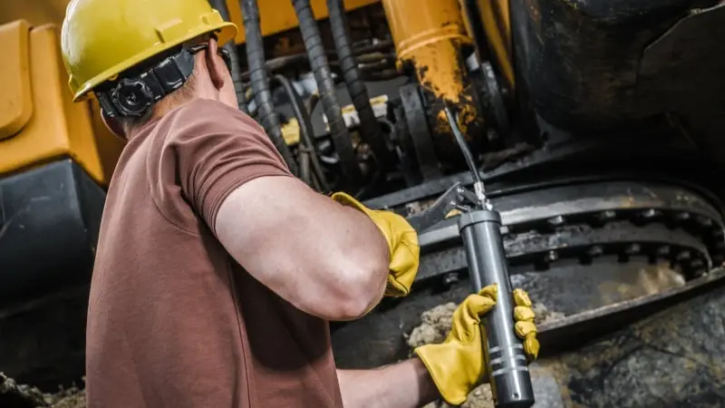 Worker in a brown shirt and yellow hard hat uses a grease gun on machinery. He wears yellow gloves. Background shows industrial equipment in Miami,FL.