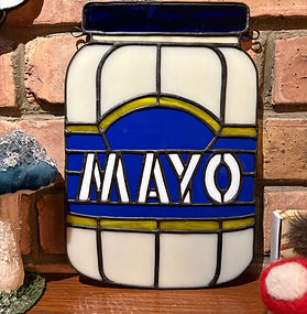 Stained glass artwork of a mayonnaise jar with blue and white labeling, displayed upright against a brick wall.