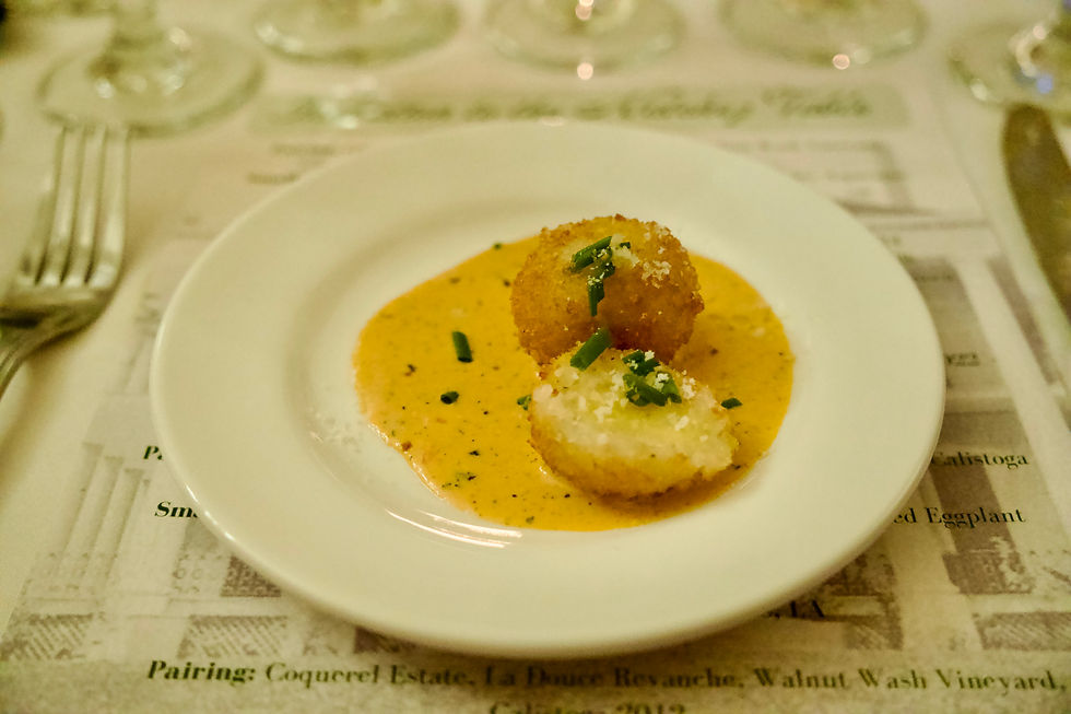 Two arancini balls on a white plate with creamy orange sauce and green garnish. Elegant table setting with glassware in background.