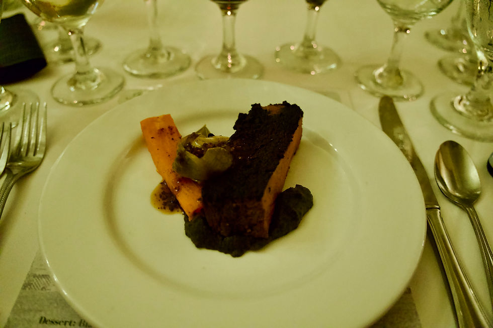 Elegant dining scene with a plated dessert, featuring dark waygu steak, caramelized carrot, sauce, and wine glasses in the background.