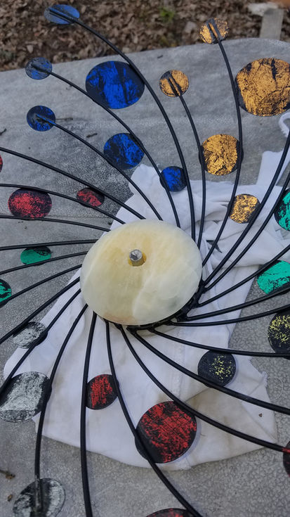 Spinning metal art with colorful glass and a central beige disc on pavement.