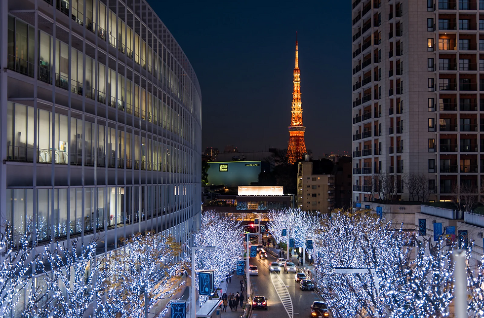 Rua iluminada com luzes brancas, prédios altos nas laterais e torre iluminada ao fundo. Atmosfera festiva e noturna em cidade movimentada.
