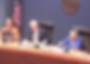Officials in a meeting sit at a wood table with laptops and papers, discussing. An American flag and city seal are visible in the background.