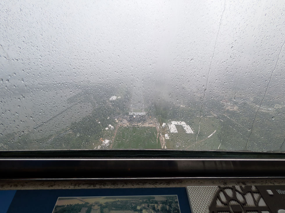 Rain-speckled window overlooking a foggy cityscape and green fields from an observation deck.