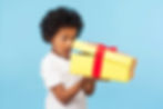 A young boy with curly hair and a surprised expression opening a yellow gift box with a red ribbon, receiving birthday presents.