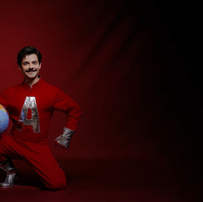 Ator vestido com roupa de superherói com um A no peito. Ele está ajoelhado, segurando um globo.