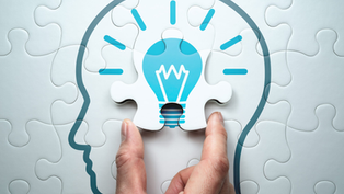 Hand placing puzzle piece with lightbulb design into head silhouette, symbolizing an idea.