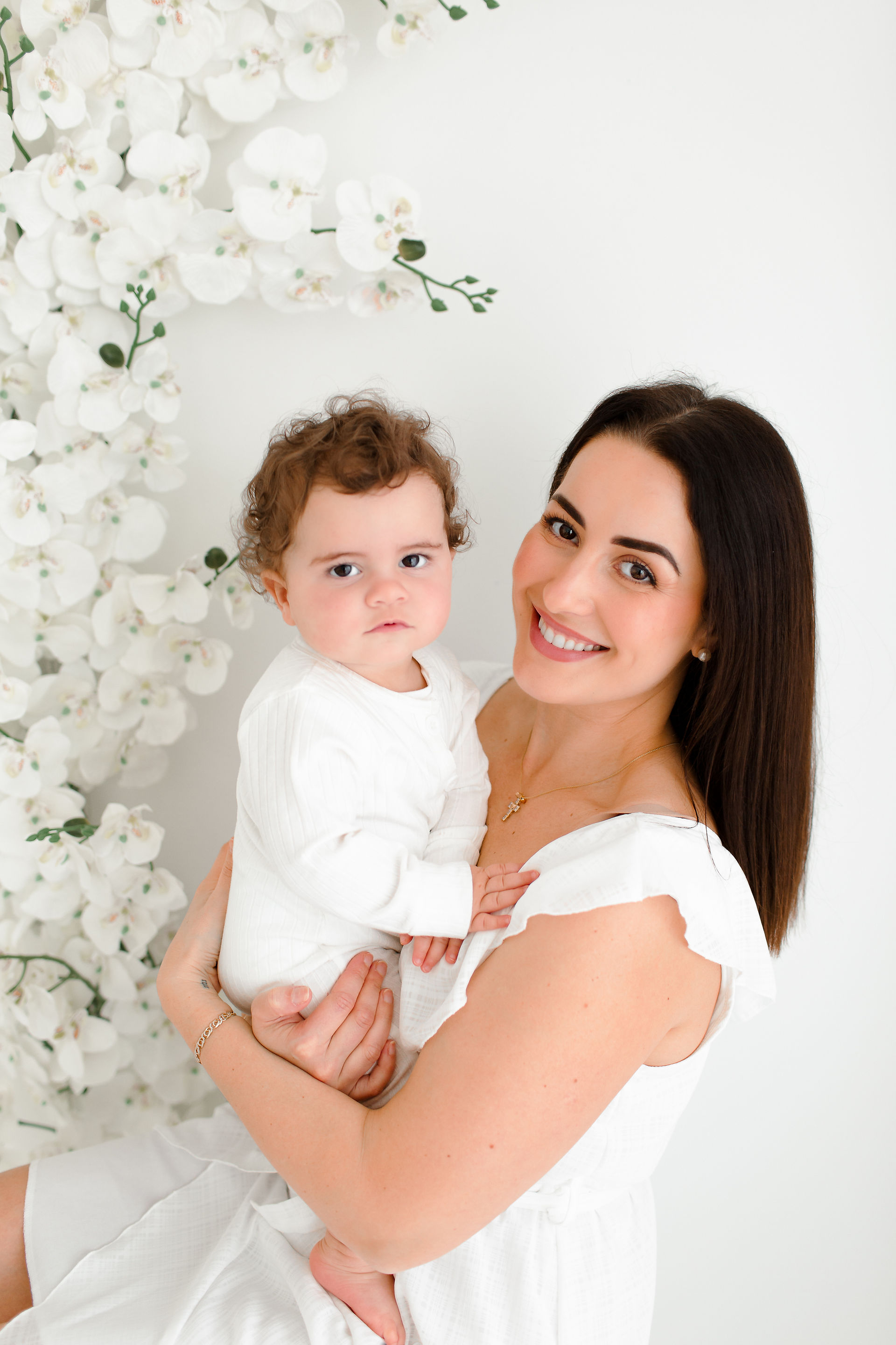 all white setting with blurred white orchids in the background, mum holding son smiling