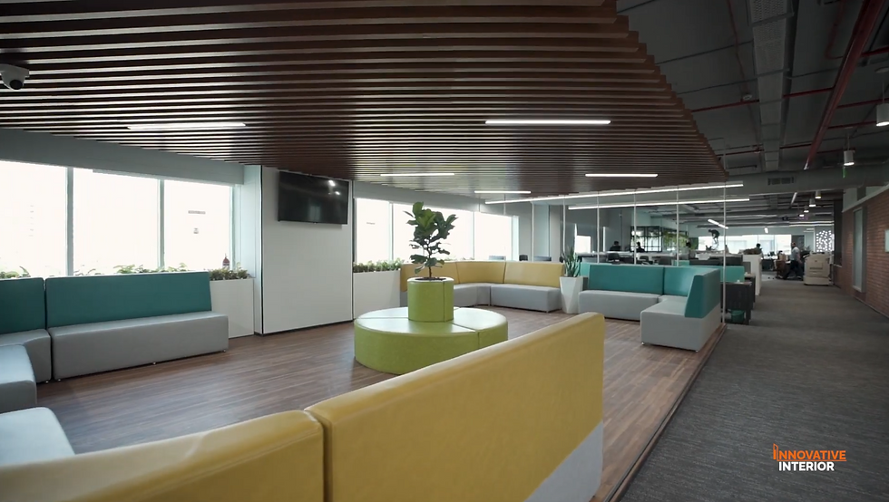 Contemporary open-plan office breakout lounge featuring colorful modular seating in yellow, teal, and grey, wood batten ceiling, indoor plants, and glass-partitioned workstations — by Innovative Interior.