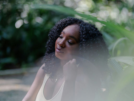 Woman with curly hair sits peacefully among lush green leaves, eyes closed in a soft, dreamy garden portrait.
