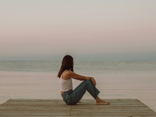 A woman sits serenely on a wooden dock, gazing out at the calm sea under a gradient sky of soft pastels.