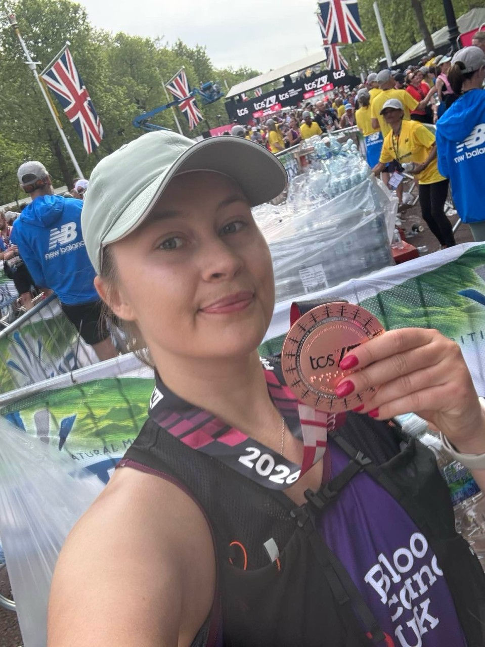 Amber Whitaker, holding her medal at the London marathon finish line. Union Jack flags, event staff, and people in background. Energetic atmosphere.
