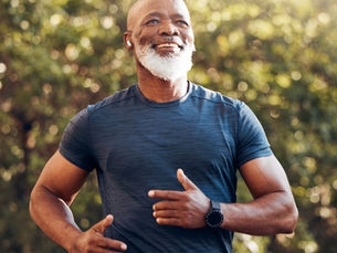 Man jogging outdoors, wearing a blue shirt and watch, smiling. Sunlit trees in the background create a warm, energetic mood.