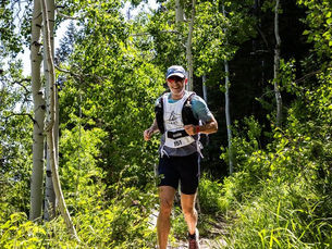 Wilson Kriegel smiles under sunlight, wearing sunglasses, cap, and race bib 151. Lush green trees surround the path. of the 29029 Trail (130km 8500ft) 