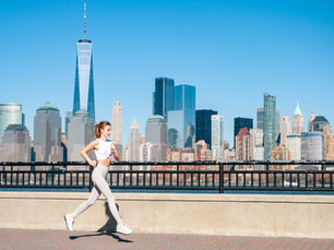 Woman jogging in white outfit along a waterfront with a city skyline in the background on a clear day.