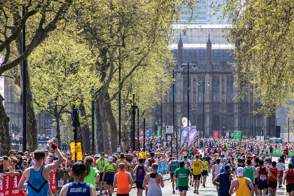 London Marathon runners on a tree-lined street approach a historic building. Colourful attire and banners; energetic and vibrant atmosphere.