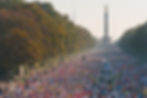 Crowd of Berlin Marathon runners race down a tree-lined street. The Victory Column in Berlin is prominent in the sunny background.