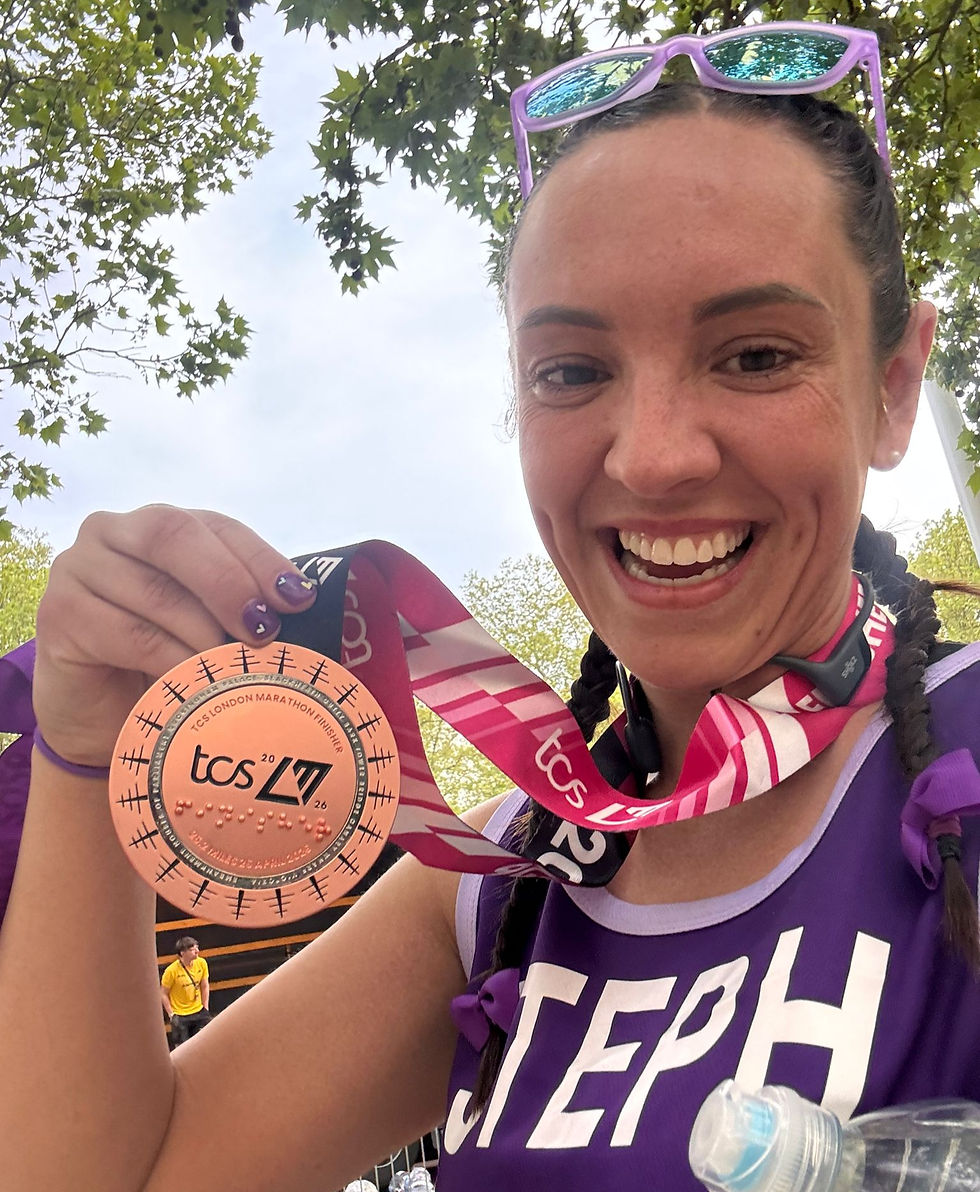 Smiling runner in purple holds a TCS London Marathon finisher medal under leafy trees, with pink ribbon and sunglasses on her head.