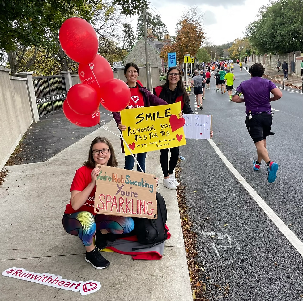 Supporters with red balloons cheer runners on a road, holding signs: Smile, You’re Not Sweating, You’re Sparkling, #Runwithheart
