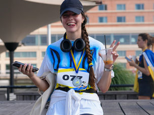 Smiling female runner in cap and medal flashes peace sign, holding phone and drink; bib 077 at a city race finish.