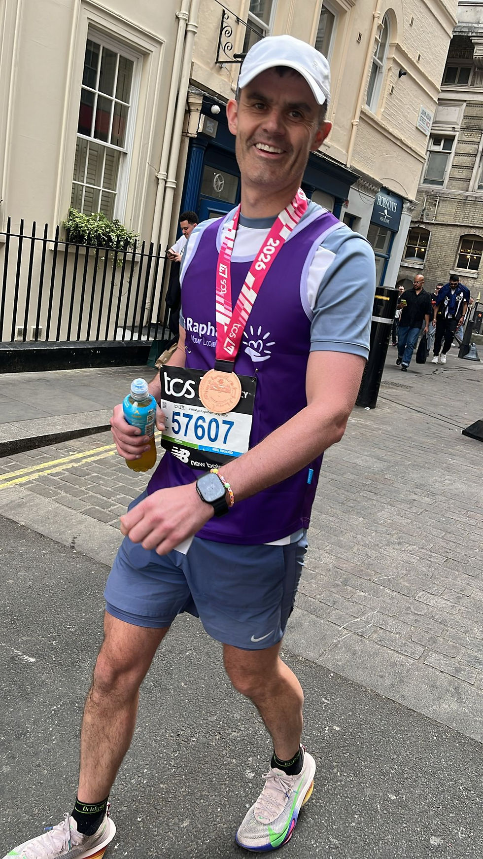 Runner in a purple vest with medal and race number 57607 walks smiling on a city street, holding a drink. Historic buildings in the background.