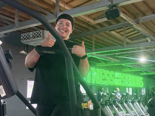 Emma Millard on her stairmaster challenge gives thumbs up on exercise machine in gym. Green neon sign in background reads "FITNESS JUST GOT SERIOUS."