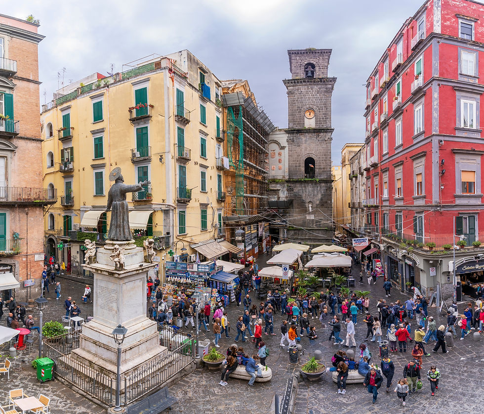 Crowded Naples piazza with a saint statue, colourful buildings, a stone clock tower, cafes, and people walking under BENVENUTI signs.
