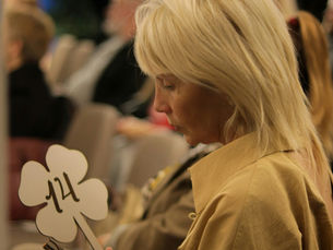Blonde woman in tan coat studies a clover-shaped sign marked 124 amid a blurred audience, looking focused and quiet.