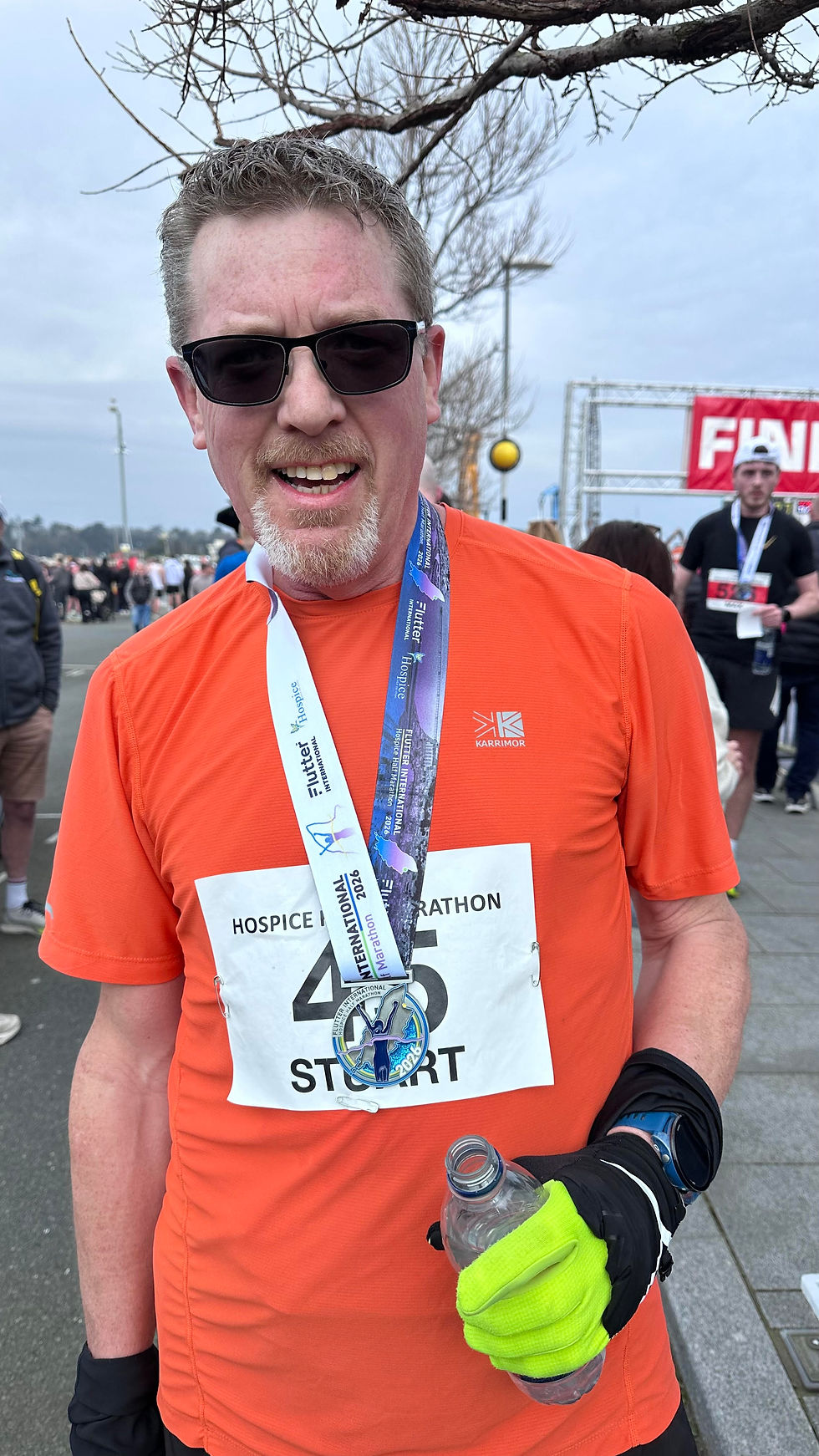 Stuart Boakesy in orange shirt with Ramsey Half marathon medal and race number 45 smiles, holding a water bottle. Background shows other runners and a "FINISH" sign.