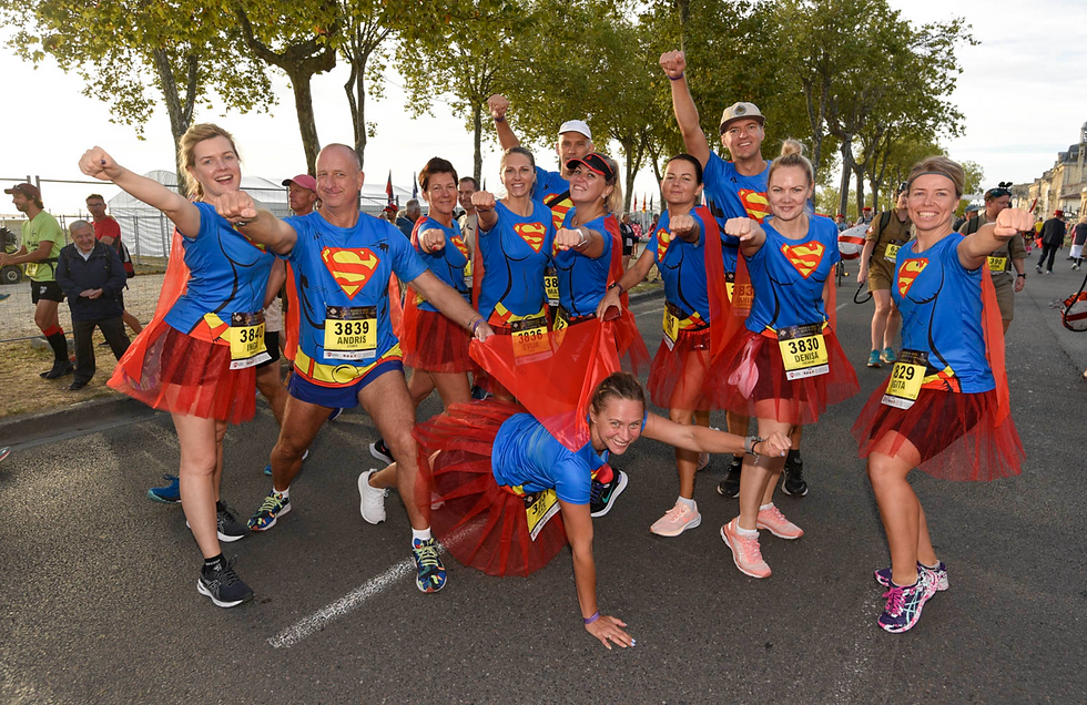 Group of runners in superhero outfits and red tutus pose playfully, raising fists. Bibs show numbers. Trees line the sunlit street. Energetic mood.