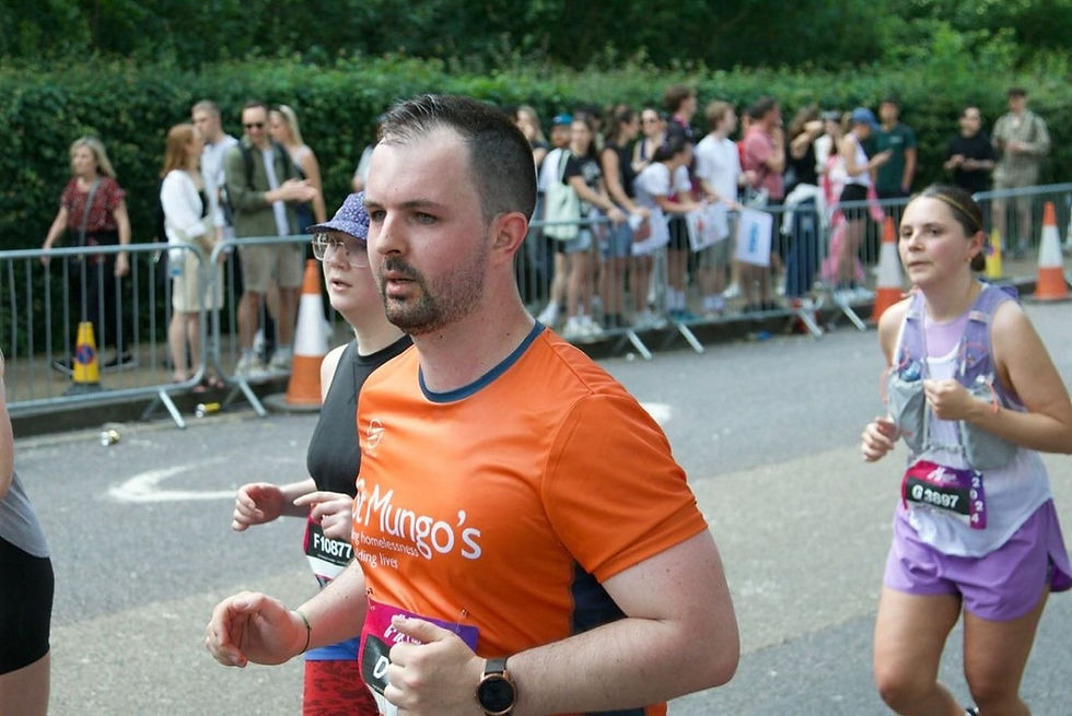 Runners in a road race pass spectators behind barriers; Matt Carpenter in orange St Mungo's shirt leads, focused.