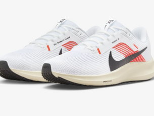 White Nike running shoes with orange and black accents, showing the text "No Human is Limited" and "Pegasus 40." Features textured sole in a studio setting.