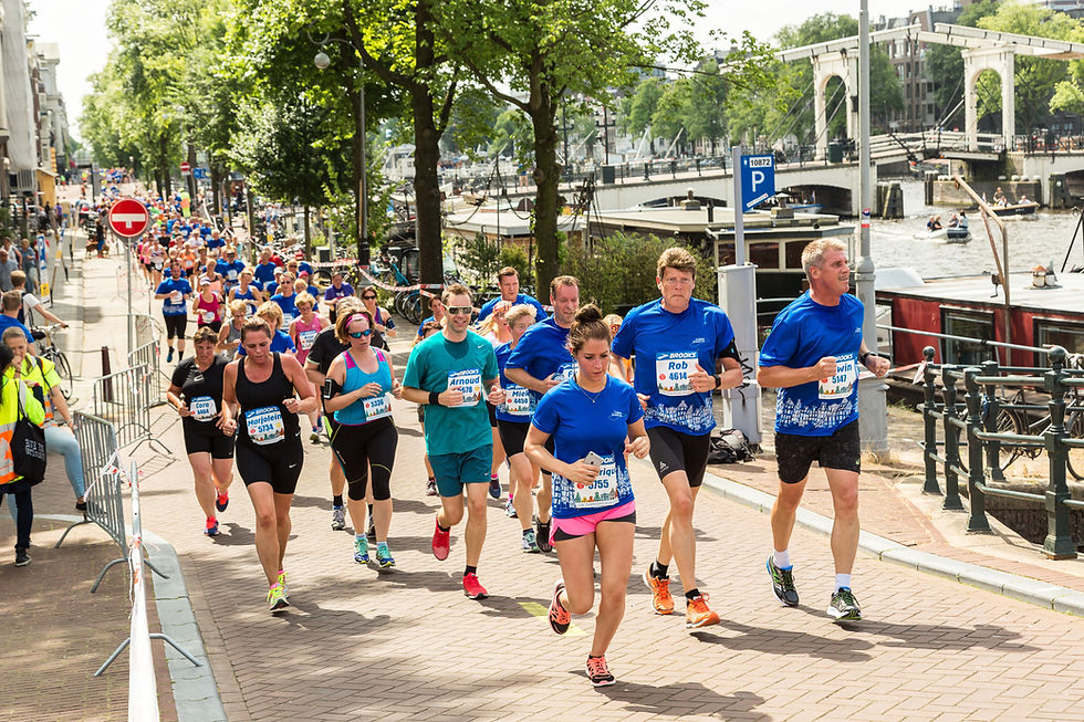 Amsterdam Marathon runners in blue shirts race along a tree-lined street beside a river, with a large bridge in the background. Mood is energetic.
