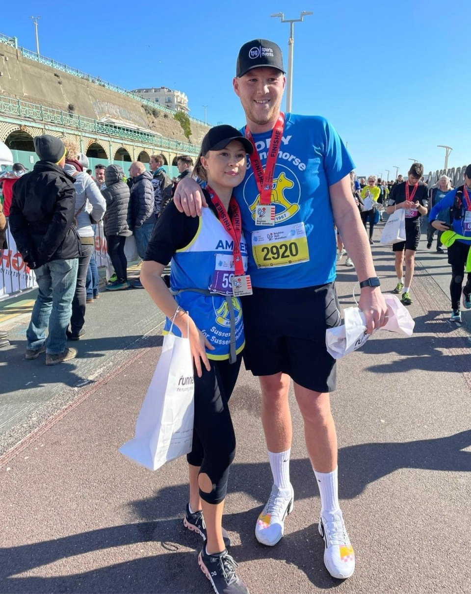 Amber Whitaker and partner pose with medals and bibs on a sunny seaside promenade, smiling after a half marathon.