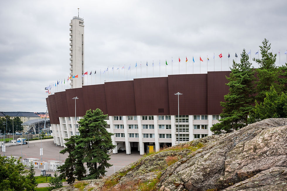 Helsinki Olympic Stadium with many flags, a tall white tower, and trees beside rocky foreground under a cloudy sky. Start and finish of the Helsinki City Half Marathon.