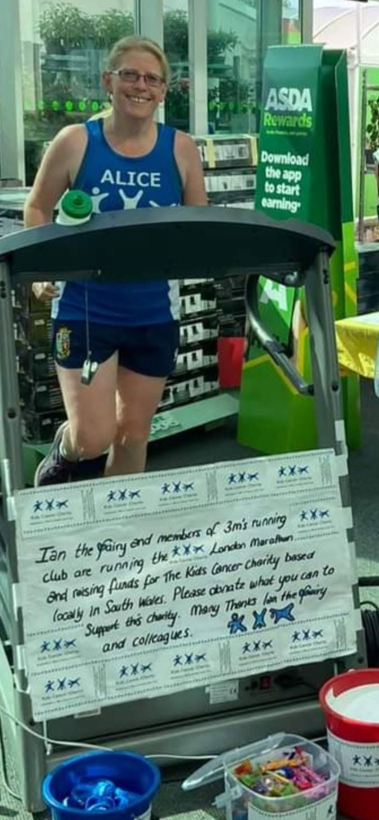 Alice Sullivan in a blue running top stands on a treadmill in an ASDA store beside a charity sign for kids cancer fundraising.