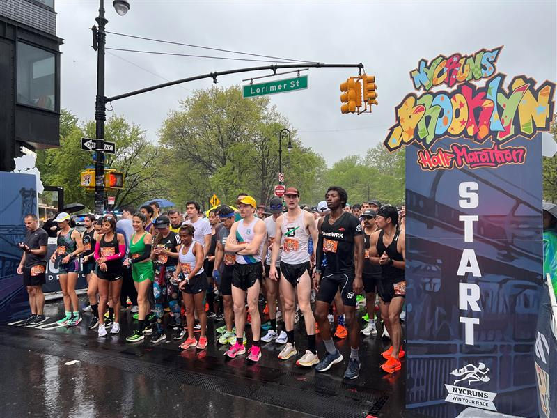 Runners gather at the start line of the Brooklyn Half Marathon on a wet street. Bright sign reads "NYCRUNS Brooklyn Half Marathon."