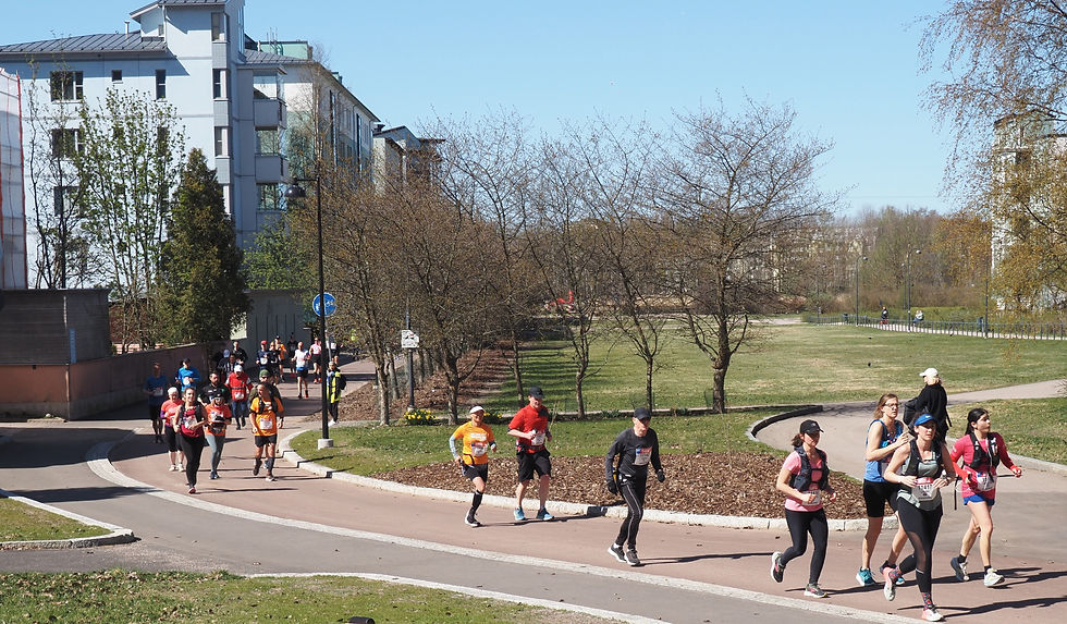 Runners with bibs jog along a sunny park path in Helsinki beside apartment buildings and leafless trees. Home of the Helsinki City Half Marathon.