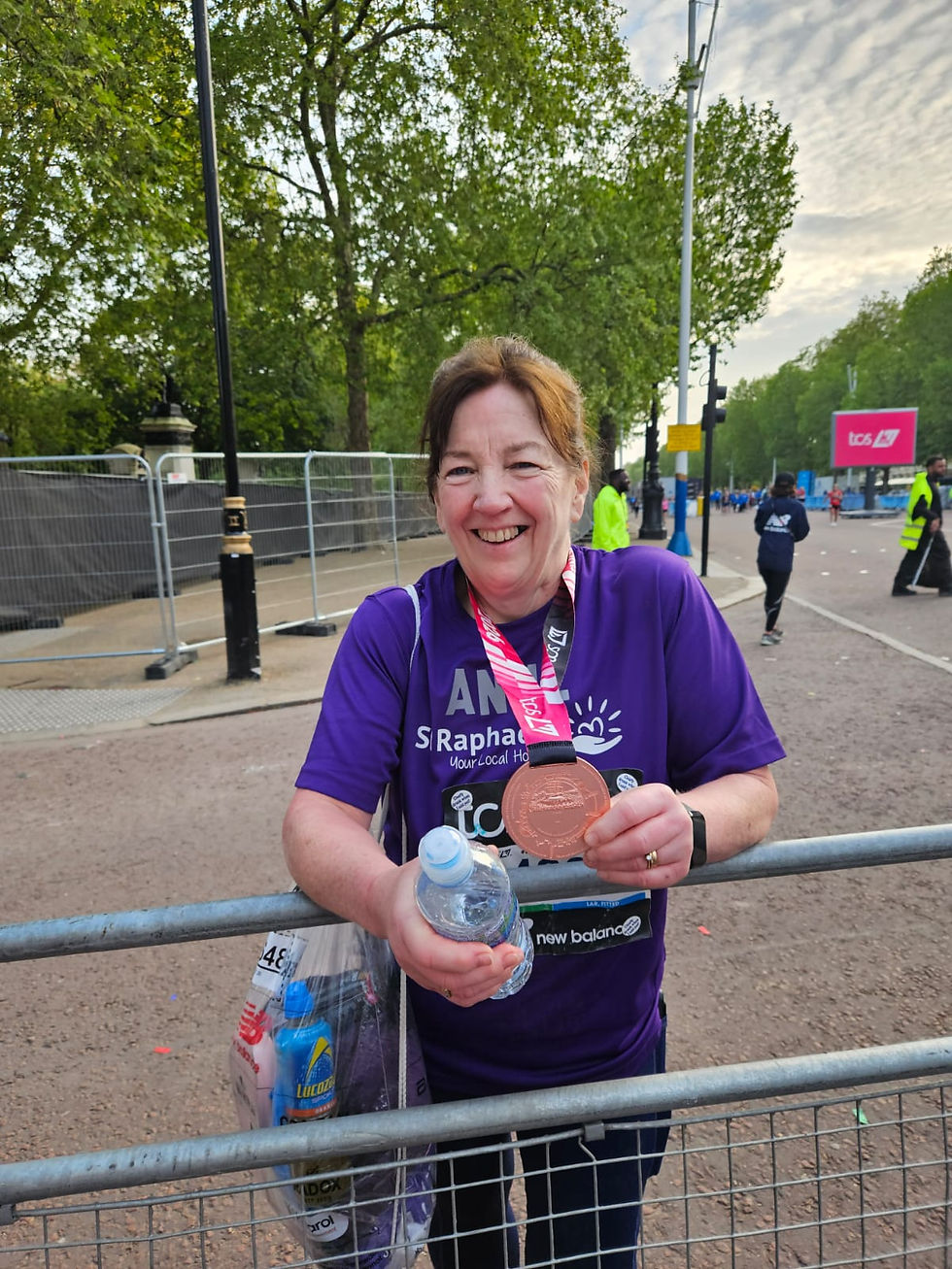 Woman in a purple shirt holds a medal and water bottle, smiling after a race. Trees and race barricades in the background.
