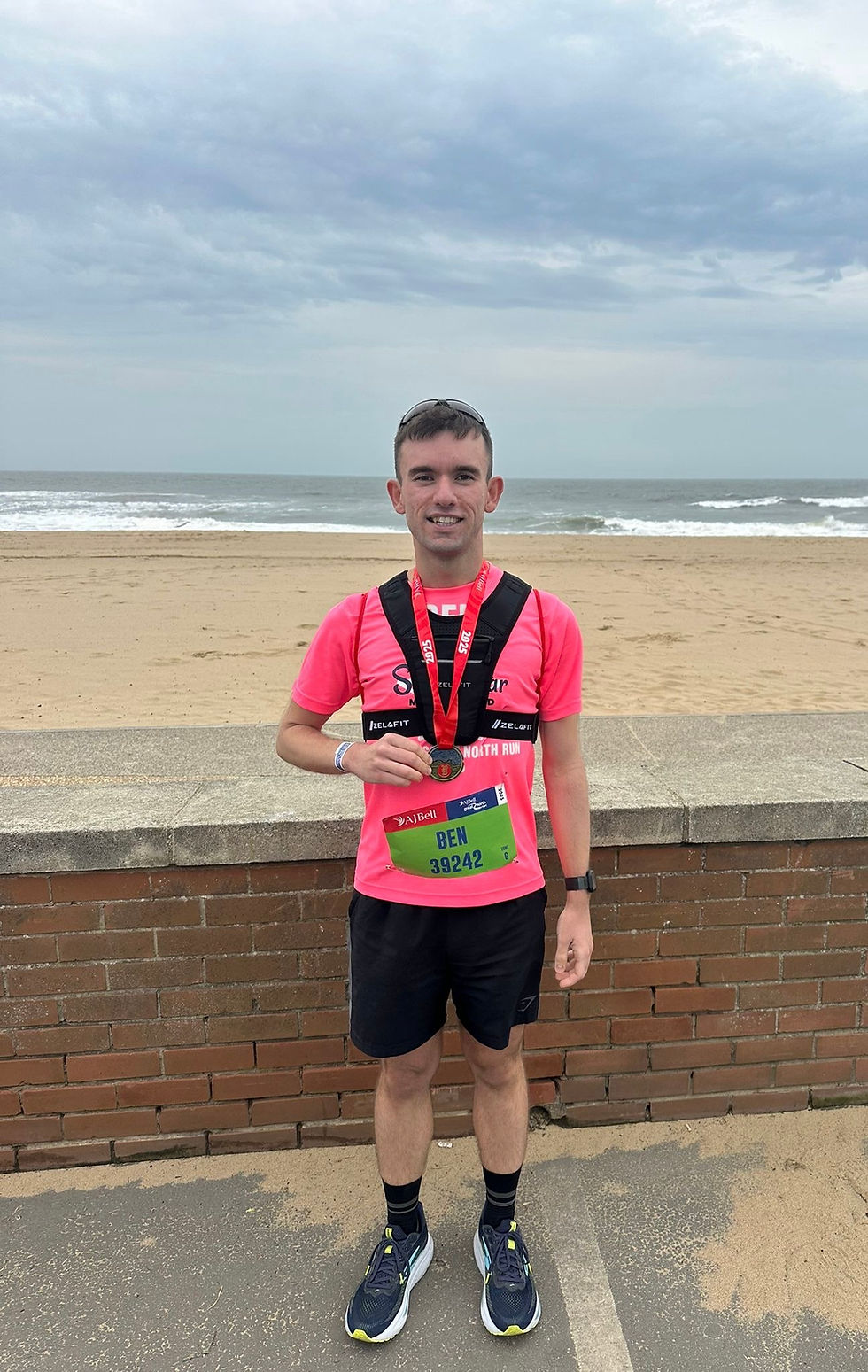 Ben Gregoriou in pink shirt with medal, bib number 39242, posing on a beach with waves and cloudy sky. Smiling, relaxed atmosphere.