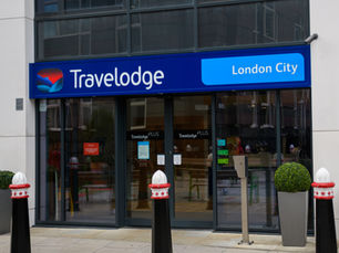 Travelodge London City entrance with blue signage. Glass doors reflect buildings. Green potted plants by the door. Urban setting.