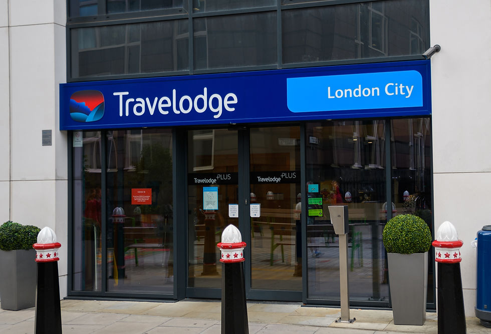 Travelodge London City entrance with blue signage. Glass doors reflect buildings. Green potted plants by the door. Urban setting.