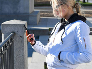Blonde woman in a white jacket checks her phone outdoors by stone steps and a railing on a sunny day.