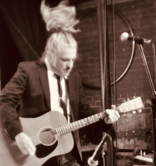 Black and white photo of The King Teen in a black suit and tie, white shirt, whipping his pony tail as he plays guitar. 