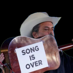 The King Teen holding up his guitar to show the back of it to the audience. A sign taped on it says SONG IS OVER.